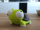 Goofy turtle figure with movable eyes sitting on a wooden desk next to a plant