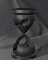 Black broken hourglass sculpture on a white desk — minimalist art sculpture close-up