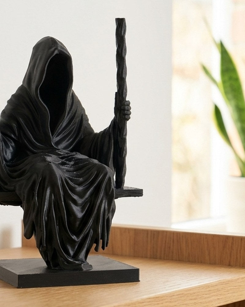 Grim reaper sculpture in matte black, hooded figure seated on a swing, front view on white background