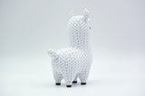 Knitted llama decorative figure styled on a wooden shelf next to a small plant and books