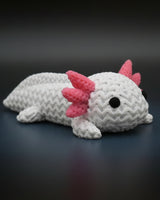 Small knitted axolotl figurine styled on a wooden shelf beside books and a plant in a Scandinavian interior