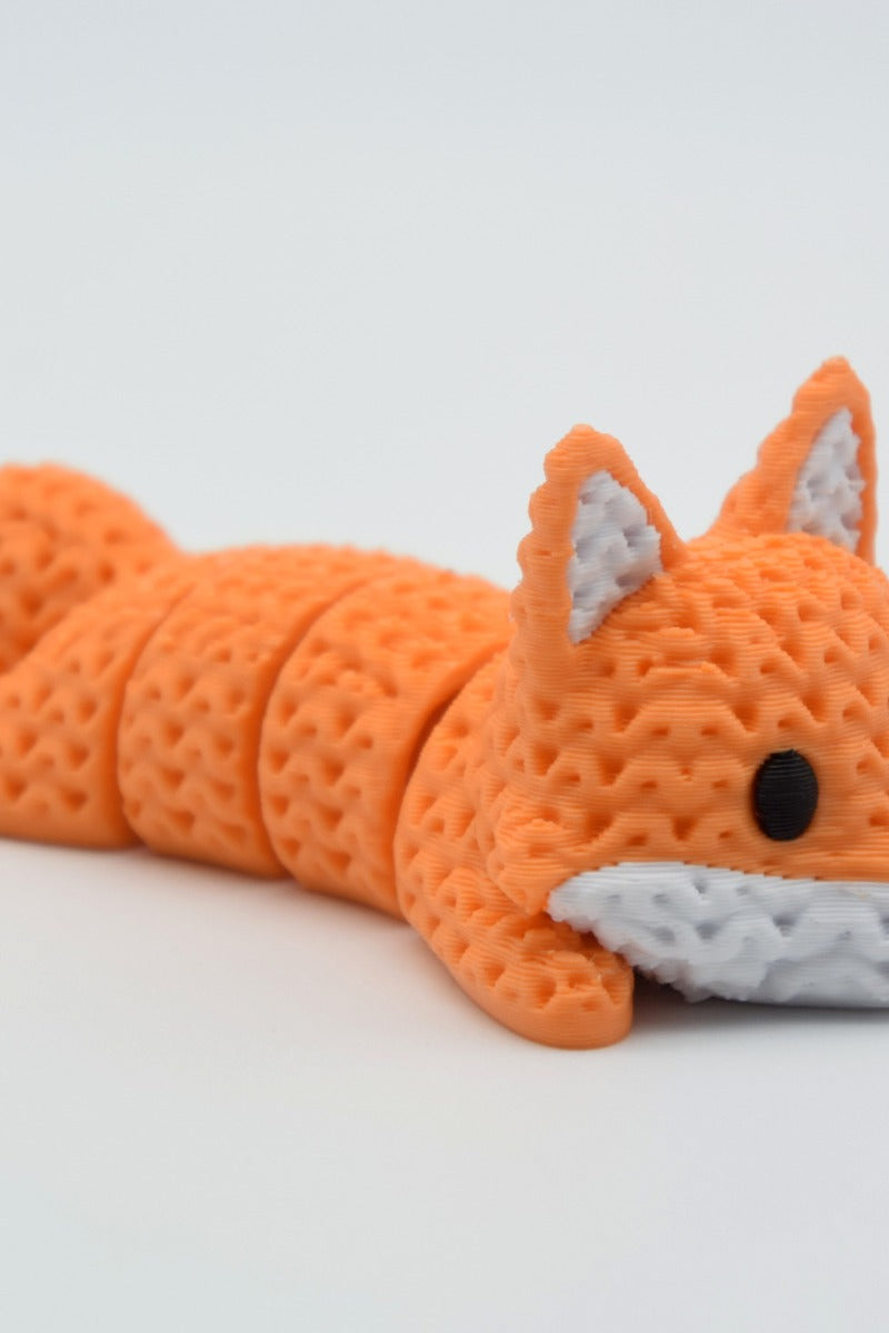 Knitted articulated fox lying on a white surface, showing its flexible segmented body and crochet-like texture