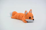 Knitted articulated fox lying on a white surface, showing its flexible segmented body and crochet-like texture
