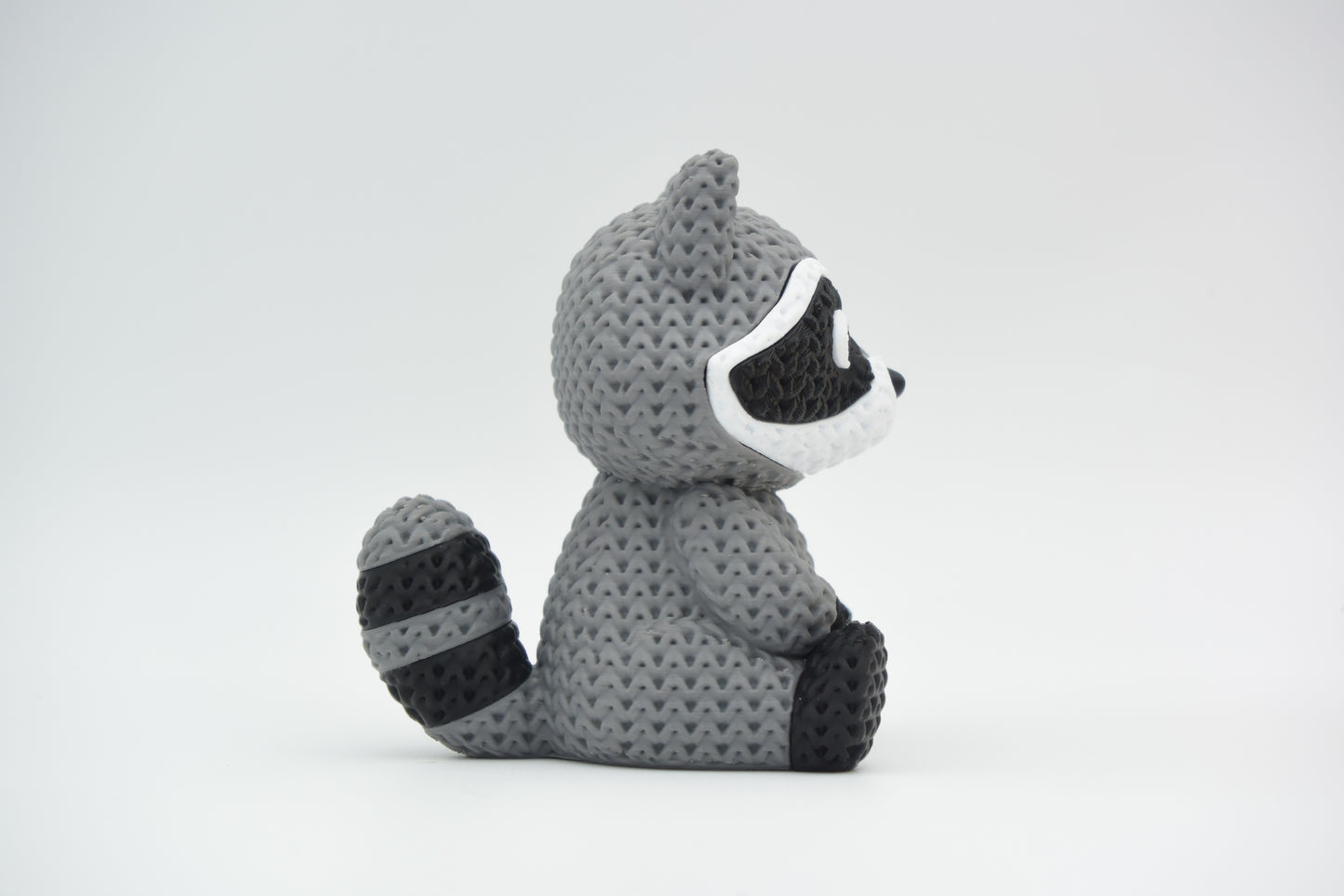 Close-up of a knitted raccoon figurine with a detailed crochet-look texture, shown against a white background