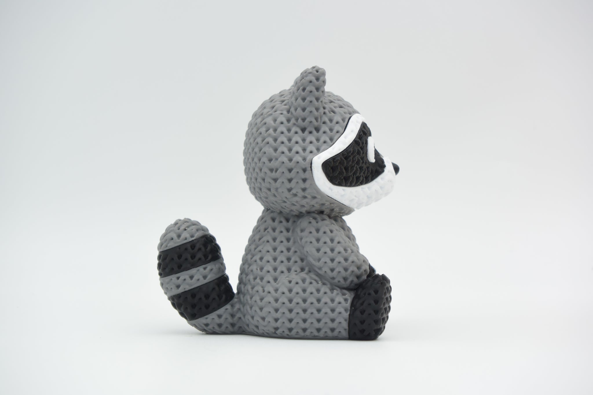 Close-up of a knitted raccoon figurine with a detailed crochet-look texture, shown against a white background