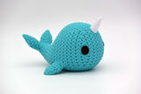 Small knitted narwhal decorative figure placed on a wooden shelf beside a candle and a few pebbles in a Nordic interior
