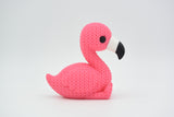 Knitted flamingo decorative figurine styled on a wooden shelf beside a small plant and neutral home decor