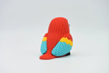 Knitted scarlet macaw parrot figurine styled on a wooden shelf beside books and a small plant