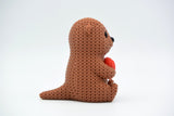 Close-up of a crochet otter figurine holding a small heart, with soft knitted texture detail, on a light surface