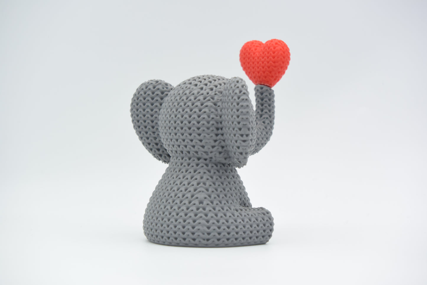 Close-up of a knitted elephant decoration blowing a heart, showing soft crochet-textured surface in warm grey
