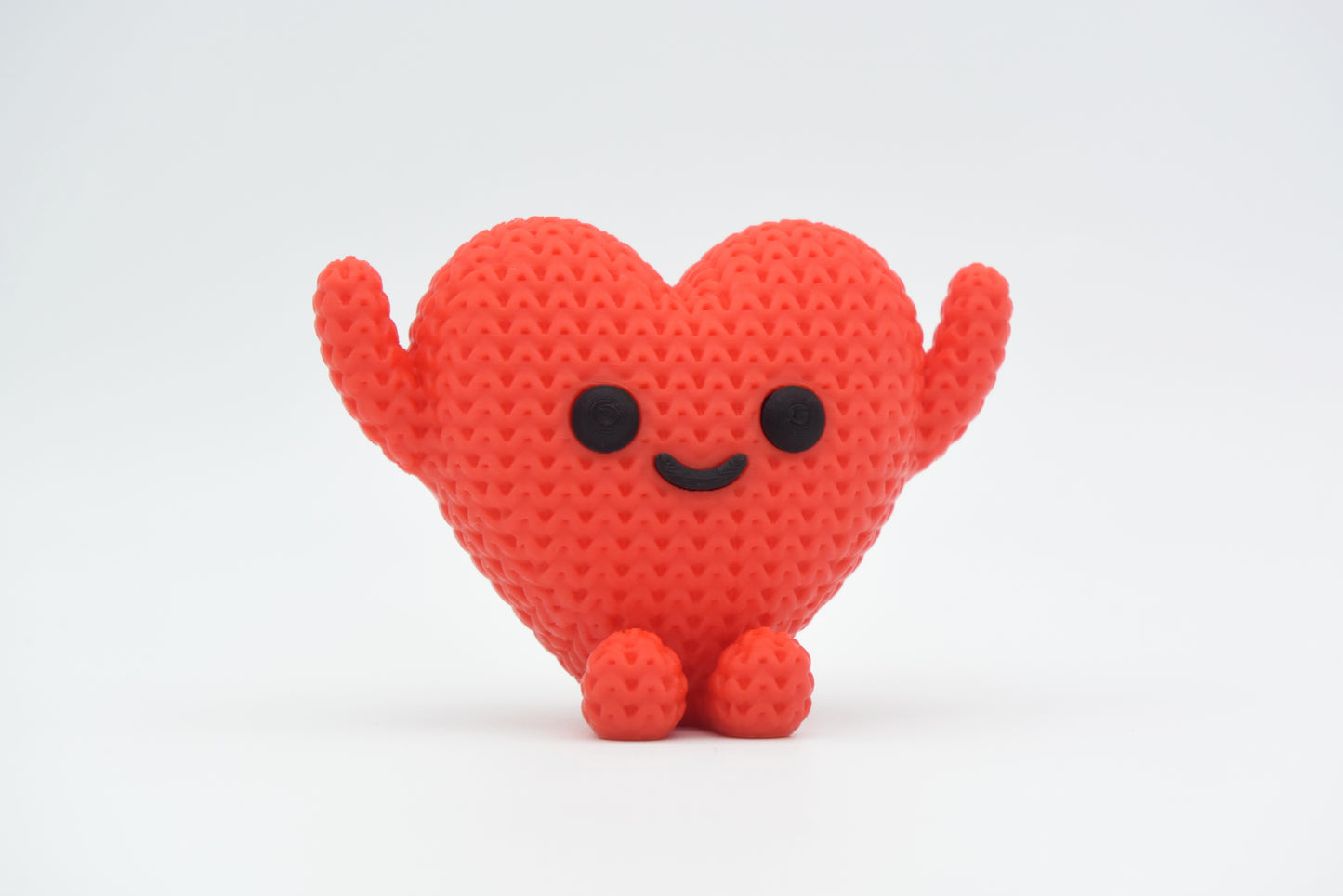 Close-up of a small knitted heart figure with a textured crochet surface and a simple character face, in red