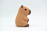 Small knitted capybara decorative figure placed on a wooden shelf beside plants and books in a Scandinavian interior